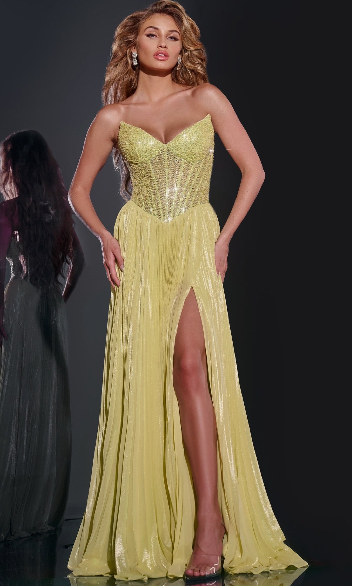A woman poses in the Jovani 43115 Long Prom Dress, featuring a strapless embellished corset bodice and a yellow floor-length skirt with a thigh-high slit, set against a dark background.